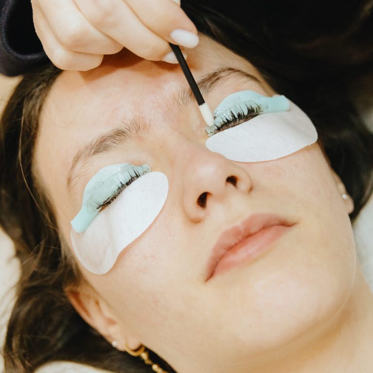 Portrait of a young unrecognizable Caucasian girl cosmetologist in a dark jacket who washes off brown paint from an eyelash on a silicone roller of her left eye using a microbrush to a teenage client lying on a cosmetology table in a home beauty salon for eyelash lifting, top side close-up view with selective focus.